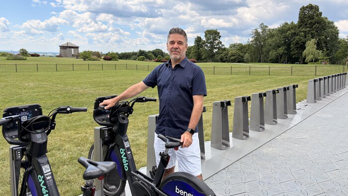 Michel de Mauraige est debout et tient un vélo inséré dans sa borne, devant le jardin Jeanne-d'Arc