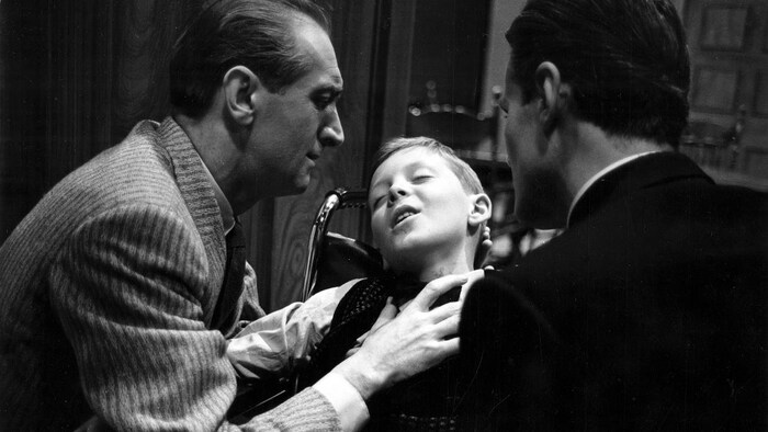 Le Dr Morell (Georges Groulx), l'air soucieux, soutient la tête du jeune Jimmy Mages (Michel Désautels), assis dans un fauteuil roulant, les yeux fermés, sous le regard de Mark Ahern (Gérard Poirier), de dos.