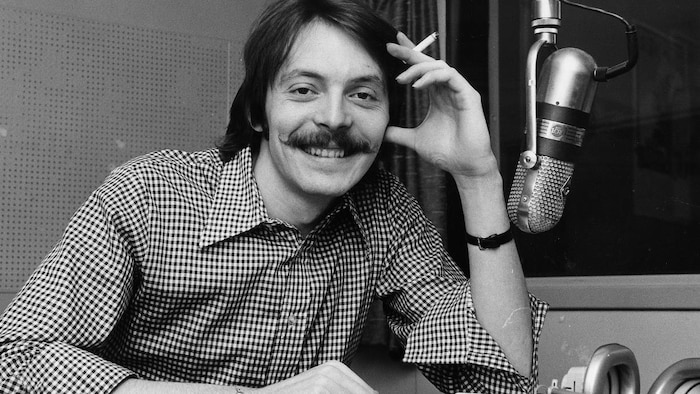 Dans un studio de radio de la station CJBC, à Toronto, l'animateur Michel Désautels, assis à une table derrière un
micro suspendu, avec une cigarette à la main.