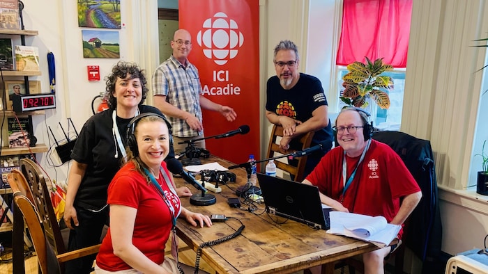 Michel Doucet et quatre employés de Radio-Canada vêtus de vêtements à l'effigie de Radio-Canada sont assis autour d'une table lors de l'enregistrement d'une émission.  Il y a des micros. 
