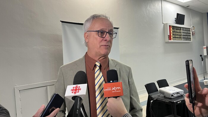Un homme mature portant des lunettes, une veste beige et une cravate rayée s'adresse à des journalistes. Plusieurs microphones, dont ceux de Radio-Canada et d'ATM, sont tendus vers lui dans une salle de conférence équipée d'un projecteur.
