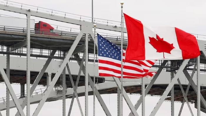 Un camion traverse un pont, avec un drapeau canadien et un drapeau américain visibles. 