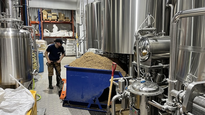 Une personne manipule un diable portant un bac près de cuves de brasserie. 