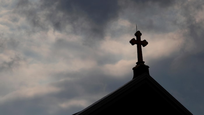 Des nuages noirs se glissent à l'arrière-plan d'une croix sur le toit d'une église catholique.