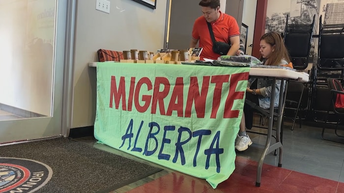 Deux personnes sont installées derrière une table de Migrante Alberta lors d’un événement communautaire. Une bannière aux couleurs de l’organisme est affichée devant la table.