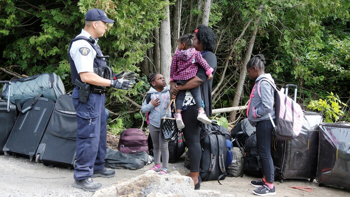 Une femme et ses trois enfants entourés de valises parlent à un agent armé.