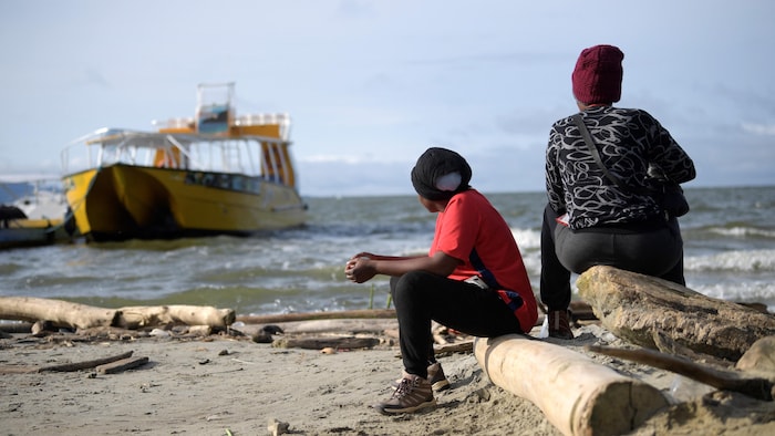 Deux personnes sont assises au bord de la mer. Elles regardent l'horizon et un bateau à quai.