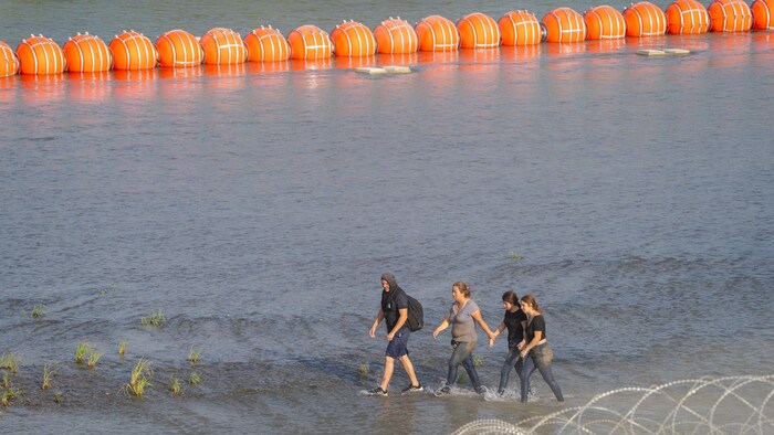 Des migrants marchent dans l'eau devant une barrière de bouées flottantes.