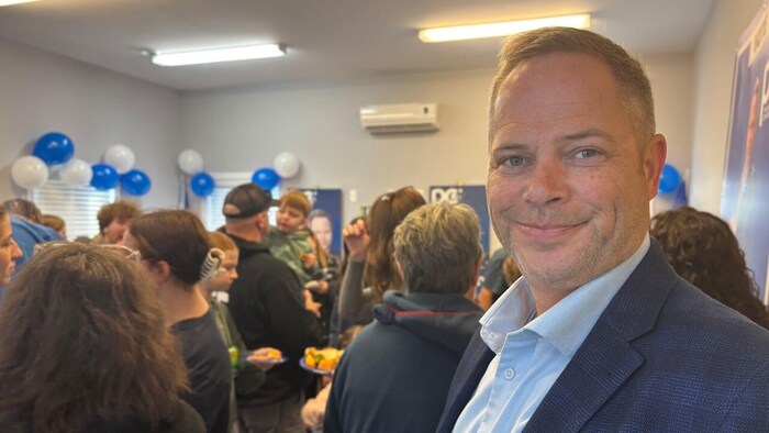 Dans une salle remplie de gens et décorée avec des ballons bleus et blancs, Mike Goosney se tourne en souriant pour la photo.
