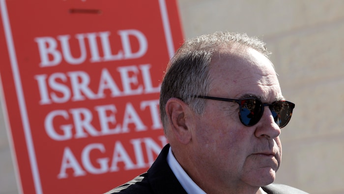 Mike Huckabee, qui porte des lunettes de soleil, est devant une affiche qui porte la mention : « Construisons à nouveau Israël de façon grandiose ».