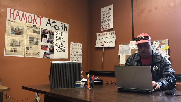 Un homme travaille à un bureau. On voit derrière lui des pancartes portant des messages à propos des logements sociaux de Hamilton.