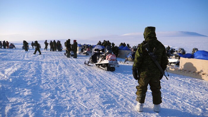 Un militaire avec son arme sur le dos regarde ses collègues et l'étendue de l'Arctique au loin.
