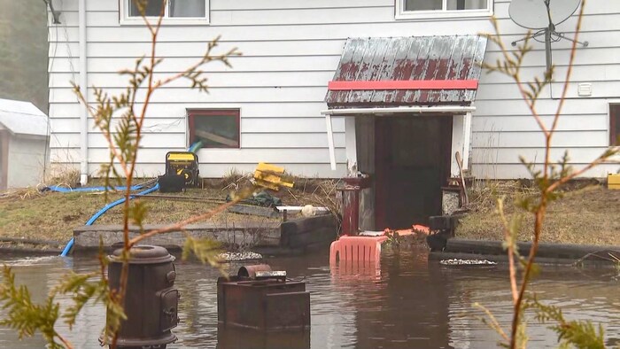 Maison au revêtement blanc partiellement inondée, avec une pompe d’évacuation active et des débris flottant dans une eau brune.