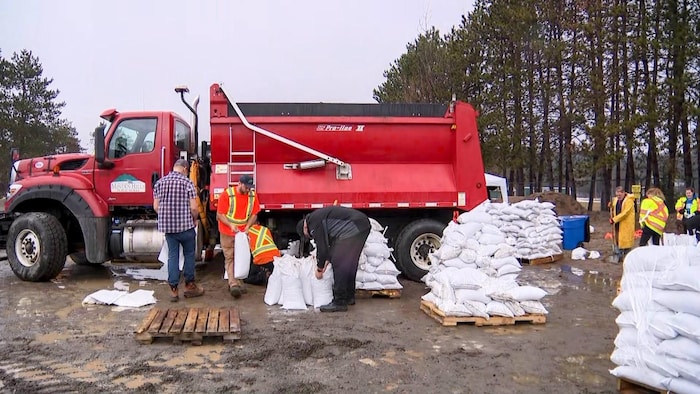 Travailleurs et bénévoles remplissant et empilant des sacs de sable près d’un camion rouge des travaux publics pour lutter contre les inondations.