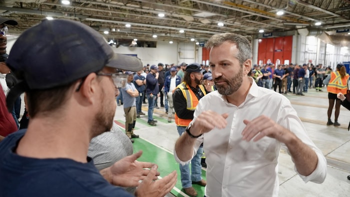Joël Lightbound discute avec un homme dans une usine. Il y a une foule de travailleurs en arrière-plan.