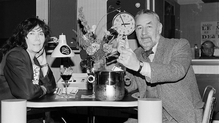Minou Petrowski en studio radio avec Philippe Noiret, un cigare à la main. Sur la petite table, un bouquet de fleurs et du vin.