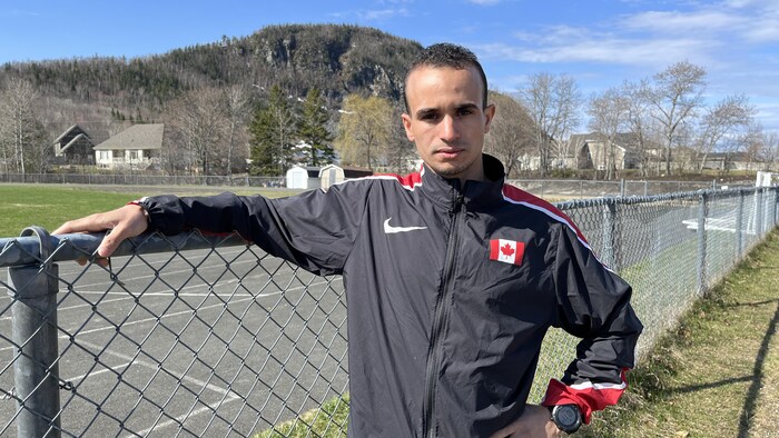 Mohamed Aagab pose pour une photo, l'air sérieux, une main sur une clôture de broche. On voit la montagne à Campbellton derrière lui.