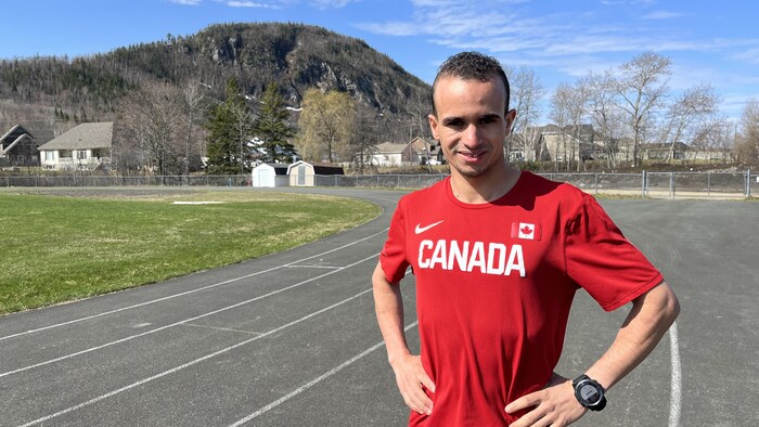 Mohamed Aagab debout sur une piste de course extérieure, les mains sur les hanches. Il porte un chandail rouge avec le mot Canada en lettres blanches. On voit la montagne à Campbellton derrière lui.