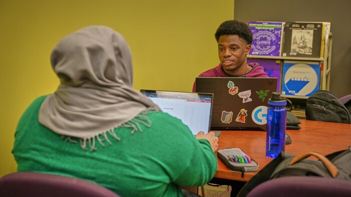 Un jeune homme est assis dans la bibliothèque avec son ordinateur portable.  Il discute avec un de ses amis. 