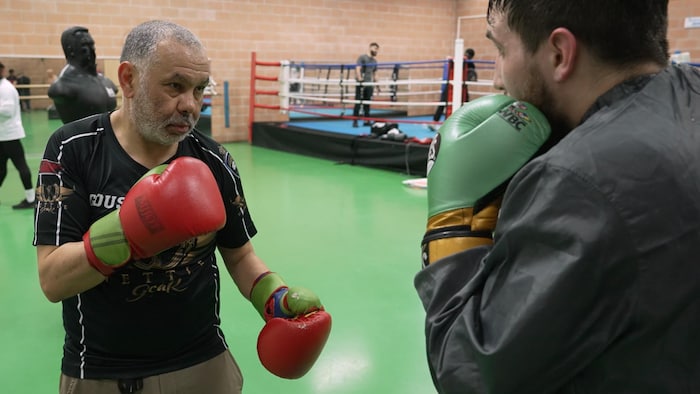 Mohammed Hanzaz, portant des gants de boxe, entraîne un jeune homme portant aussi des gants de boxe dans un gymnase, avec un ring à l'arrière-plan.