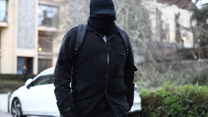 Un homme, habillé en noir, qui dissimule son visage à l'aide d'une tuque, de lunettes et d'un cache-cou.
