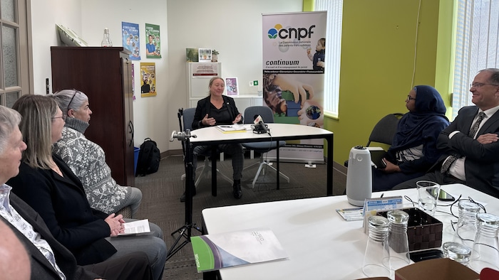 Une femme assise derrière un bureau, parle à des gens assis qui l'écoutent.