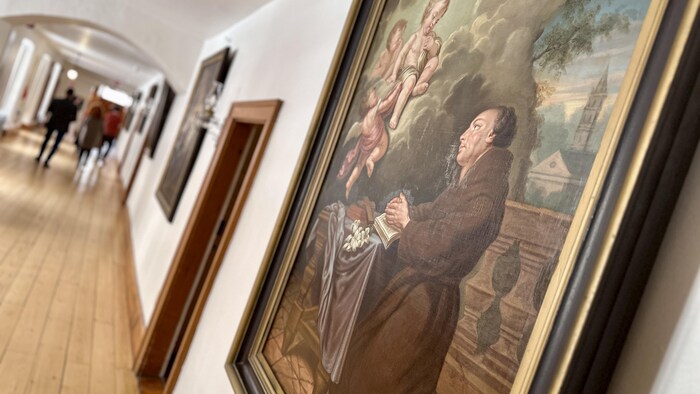 Un couloir du monastère, avec une peinture religieuse au mur. Sur celle-ci, on voit un prêtre qui prie et trois anges.