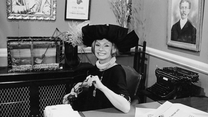 Monique Mercure, dans un costume des années 1920, est assise à un bureau, souriante.
