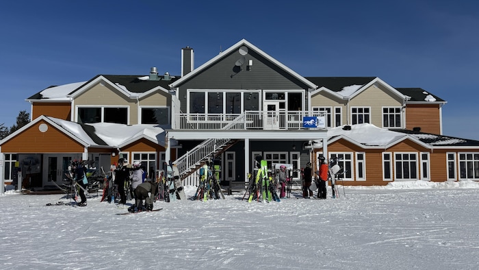Cette photo présente le chalet principal d'une station de ski sous un ciel bleu dégagé. Au premier plan, de nombreux skis et planches à neige sont appuyés devant le bâtiment, où circulent quelques personnes en tenue de sport hivernal.