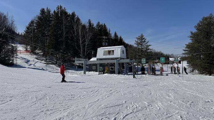 La photo montre la base d'un télésiège dans une station de ski par temps clair. Un skieur en manteau rouge descend une pente enneigée au premier plan, tandis que d'autres personnes attendent près des portillons d'accès devant la forêt.