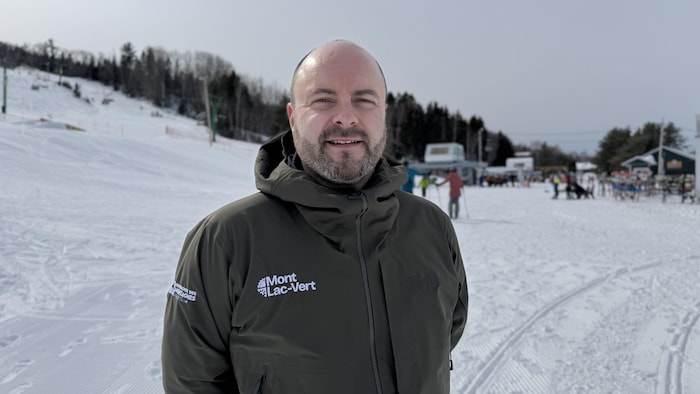 Un homme barbu et souriant pose à l'extérieur, vêtu d'un manteau de ski vert olive arborant le logo « Mont Lac-Vert ». En arrière-plan, on aperçoit une station de ski enneigée avec des skieurs, des remontées et une forêt.