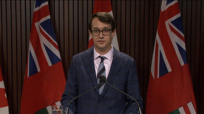 Isang lalaki na nakasalamin nakatayo sa podium habang may watawat ng Ontario sa background.