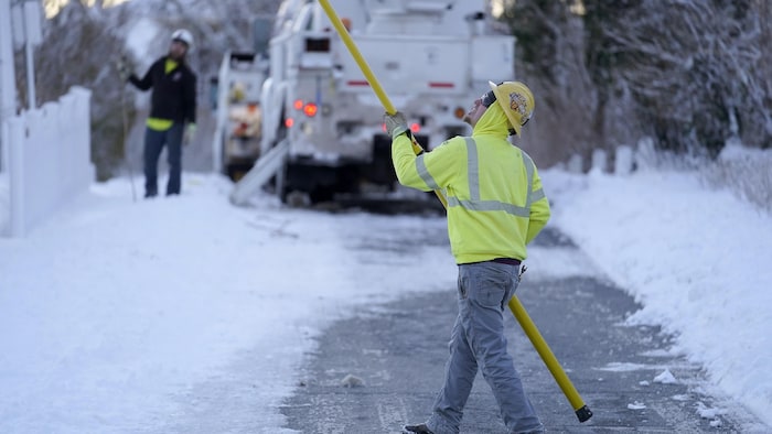 Un monteur de lignes en veste de sécurité jaune marche sur une chaussée enneigée en tenant un grand bâton et en levant la tête vers les lignes de transmission électrique.
