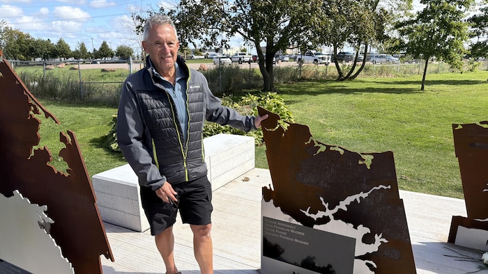 Dans un parc, Jean-Paul Arsenault pose, une main sur la sculpture en métal sur laquelle apparaît le nom de sa mère.