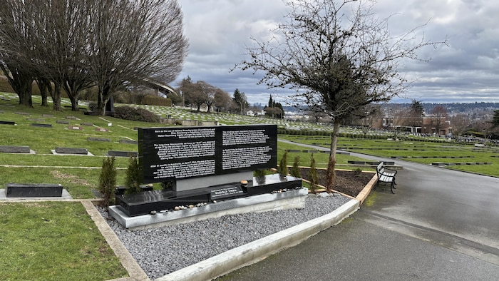 Un monument dans un cimetière, avec plusieurs arbres et un chemin en asphalte. 