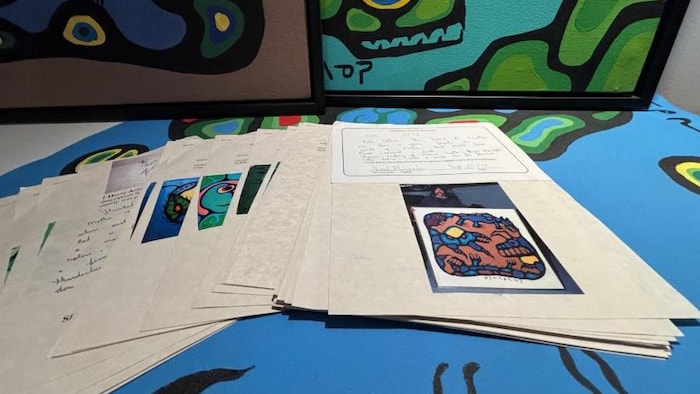 Des documents et des photographies sont disposés sur une toile, devant plusieurs tableaux aux couleurs vives.