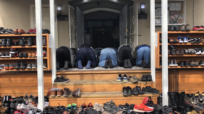 Une dizaine d'hommes à genou de dos débordent de la salle de prière. Derrière eux se trouve une cinquantaine de paires de chaussures posées à l'entrée.
