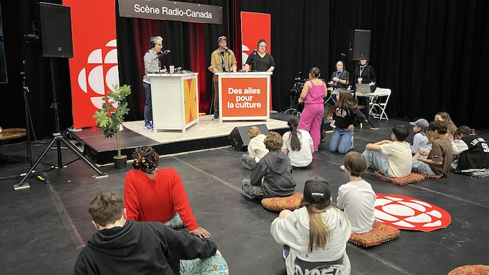 Mathieu Dugal et ses deux invités sur la scène Radio-Canada, avec des enfants assis devant en train de les écouter.