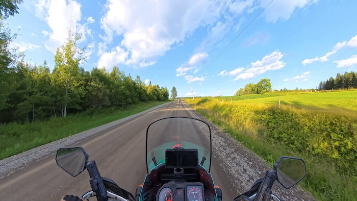 Un paysage champêtre se dessine au loin, du point de vue d'un motocycliste dont le guidon et l'avant de la moto sont dans l'image.