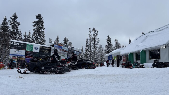 Une photo en plan large prise en hiver montre un relais de motoneigistes avec un bâtiment couvert de neige à droite. Plusieurs motoneiges sont garées devant des panneaux d'affichage. Des hauts conifères enneigés encadrent la scène.