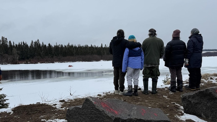Cinq personnes sont debout, de dos, sur le bord d'une rivière partiellement gelée. Une motoneige est stationnée sur la banquise au loin.