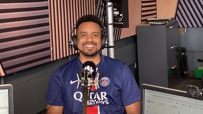 Portrait d'un homme souriant dans un studio de radio, portant un casque audio et un maillot bleu du Paris Saint-Germain.