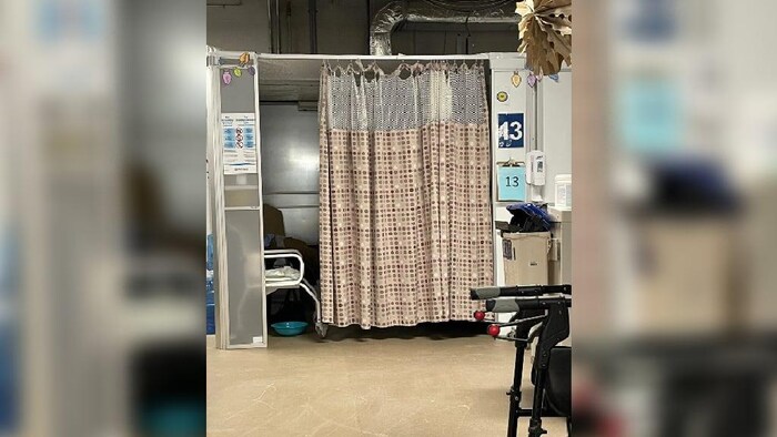 Un cubicule avec le numéro 13 où est accroché un rideau brun entrouvert, qui laisse apercevoir un pot de chambre sur le sol, sous une chaise.