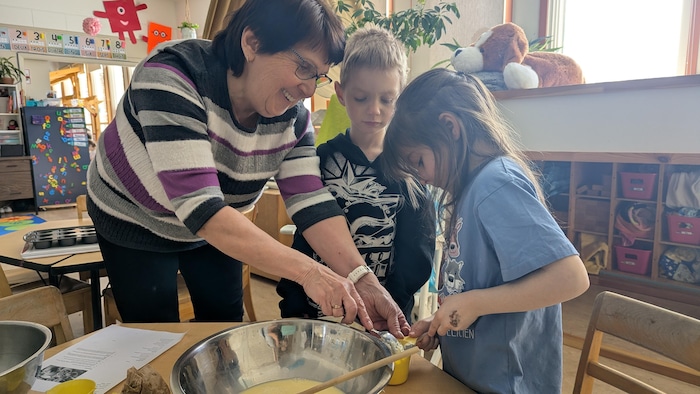 Tout sourire, la bénévole aide une fillette à brasser un mélange, sous le regard d'un garçon.