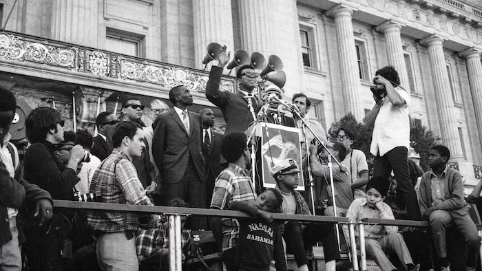 Muhammad Ali devant un pupitre qui prononce un discours entouré d'une vingtaine de personnes. 
