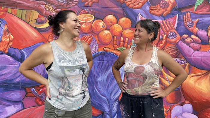 Deux femmes rient ensemble derriére elles, une murale colorée. Il s'agit des artistes Alejandra Zeme (à gauche) et Marila Tarabay, fondatrices du collectif argentin Nereidas R., qui a 20 ans en 2024. Ce sont les muralistes qui ont créé « Guardiennes des sémences » à Montréal. 