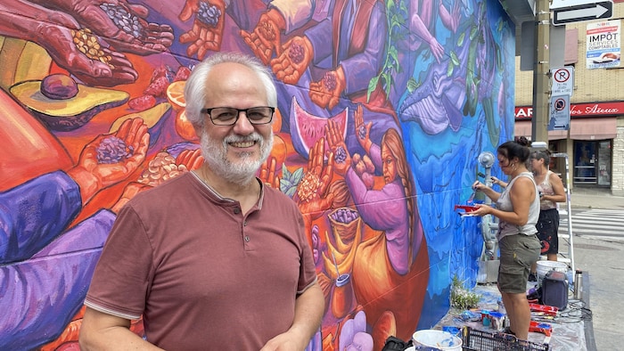 Un homme souriant à la caméra devant une murale.