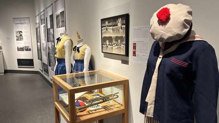 Plusieurs mannequins vêtus de costumes historiques, dont une tenue de style marin avec béret à pompon au premier plan et deux robes traditionnelles à corset à l’arrière. Entre eux, une vitrine expose une trompette ancienne et divers insignes.