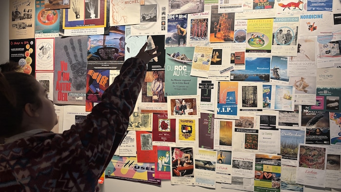 Vue de dos d'une personne levant la main pour pointer un document sur un grand mur entièrement recouvert d'affiches, de photographies d'archives et de dépliants d'expositions passées.