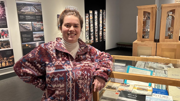 Portrait à mi-corps d’une femme souriante, la main appuyée sur le rebord d’une vitrine en bois contenant des livres et documents d’archives. En arrière-plan se trouvent des panneaux documentaires muraux et des présentoirs d'exposition.